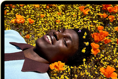 MacBook Neo, color cítrico, persona sonriente tumbada en un campo de flores en la pantalla