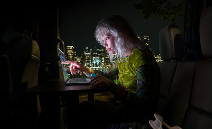 Una persona sentada en el asiento trasero de un furgón, la puerta del pasajero abierta muestra el horizonte de una ciudad por la noche, MacBook Pro con una pantalla PROLED XDR sobre una mesa de trabajo, la persona está iluminada por la pantalla mientras utiliza el control táctil para editar un vídeo en DaVinci Resolve Studio